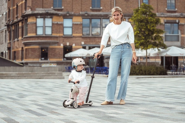 Młoda kobieta prowadzi dziecko w jeździku Scoot And Ride Highwaykick 1 Push and Go z rączką do prowadzenia Ash