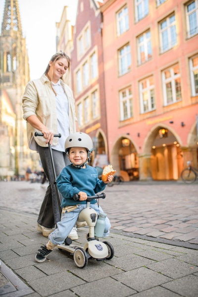 Młoda kobieta prowadzi dziecko w jeździku Scoot And Ride Highwaykick 1 Push and Go z rączką do prowadzenia Ash