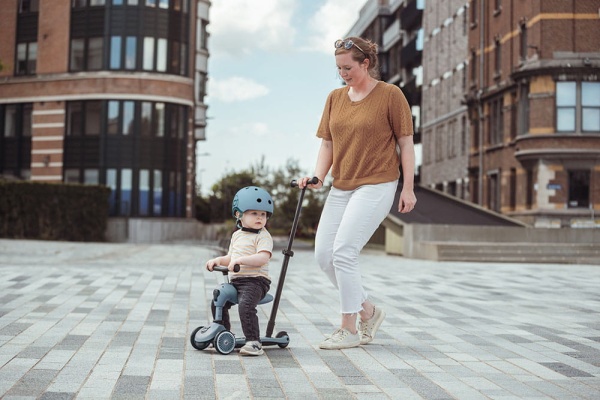 Młoda kobieta prowadzi dziecko w jeździku Scoot And Ride Highwaykick 1 Push and Go z rączką do prowadzenia Steel