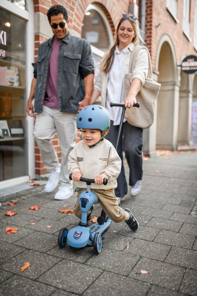 Młodzi rodzice prowadzą dziecko w jeździku Scoot And Ride Highwaykick 1 Push and Go z rączką do prowadzenia Steel